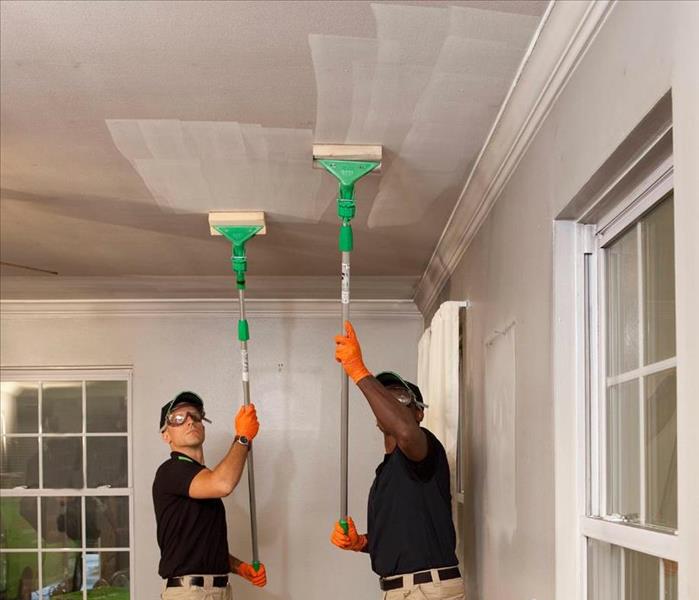 Two employees male with orange gloves using ceiling sweepers to wipe off soot in living room. SERVPRO logo on bottom right